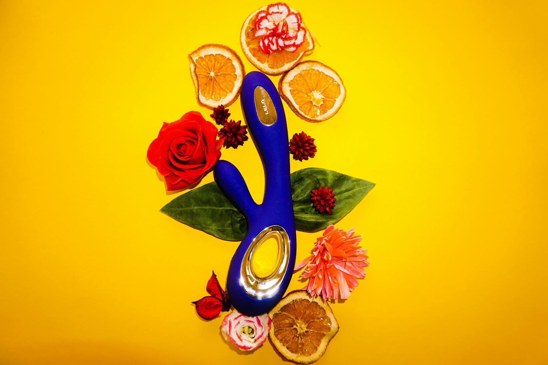 Vibrador azul cercado por flores e rodelas de laranja em fundo amarelo vibrante